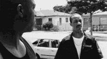 a black and white photo of two men talking in front of a car .
