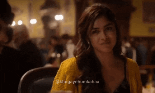 a woman in a yellow sweater is sitting at a table in a restaurant and looking at the camera .
