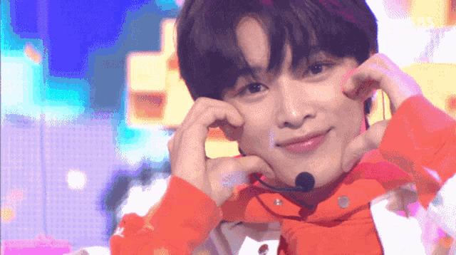 a young man wearing a red jacket and a microphone is making a heart shape with his hands