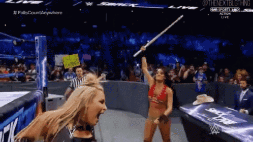 a woman in a bikini is holding a stick in a wrestling match while a referee looks on .