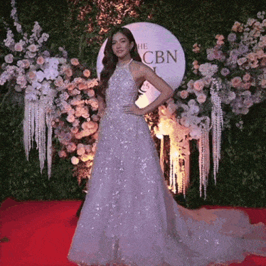 a woman in a long purple dress stands on a red carpet in front of a sign that says the cbn