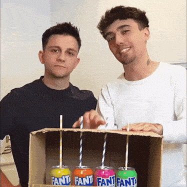 two men are standing next to a cardboard box with four fanta cans in it