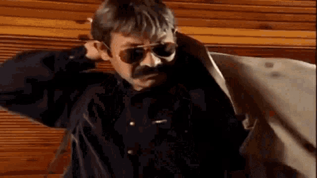 a man with a mustache wearing sunglasses and a jacket is standing in front of a wooden wall .