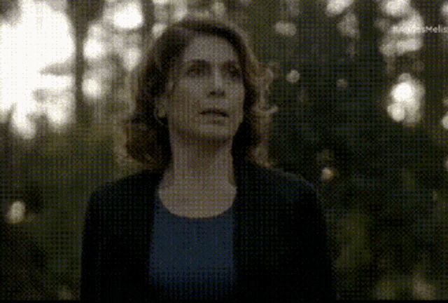 a woman in a blue shirt and a black jacket is standing in a forest looking up .