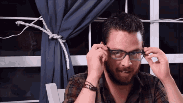 a man adjusts his glasses in front of a window with a blue curtain