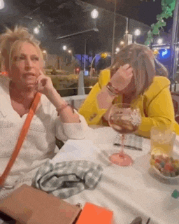 two women are sitting at a table with a glass of wine . one of the women is wearing a yellow jacket .