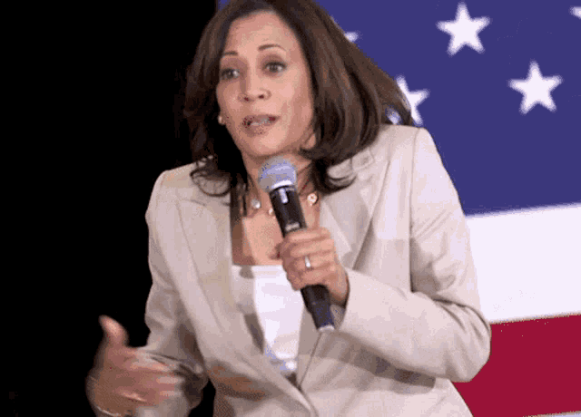 a woman in a tan suit is holding a microphone in front of an american flag