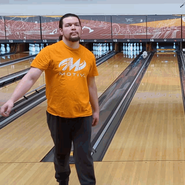 a man wearing an orange shirt that says motiv is standing in a bowling alley