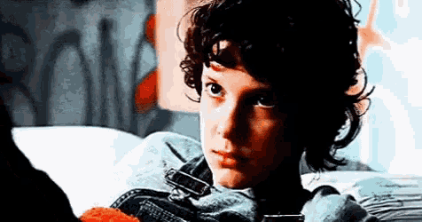 a young boy with curly hair is sitting on a bed holding a hamburger .