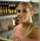a woman in a pink shirt is smiling in front of a shelf with bottles on it .