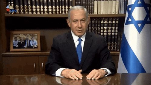a man in a suit and tie sits at a desk in front of a flag with a star of david on it