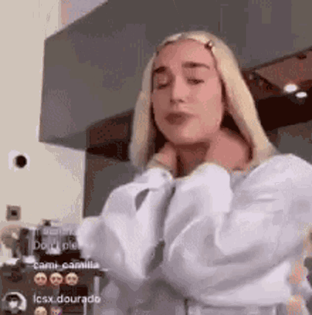 a woman in a white shirt is standing in a kitchen with her hands on her neck .
