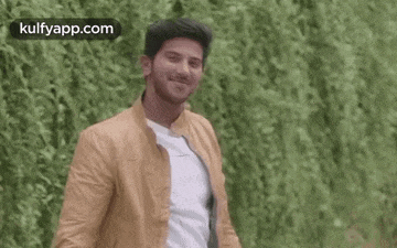 a man in a brown jacket and white shirt is smiling while standing in front of a green bush .