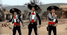 three men in mariachi costumes are standing in a desert holding guns .