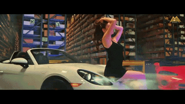 a woman in a black dress is standing next to a white car in a warehouse with the letter k on the shelves