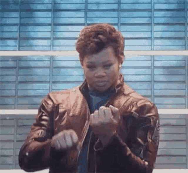 a woman in a leather jacket is making a fist with her hands in front of a brick wall .