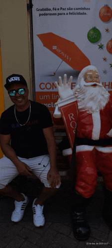 a man wearing a black vans hat sits next to a santa statue