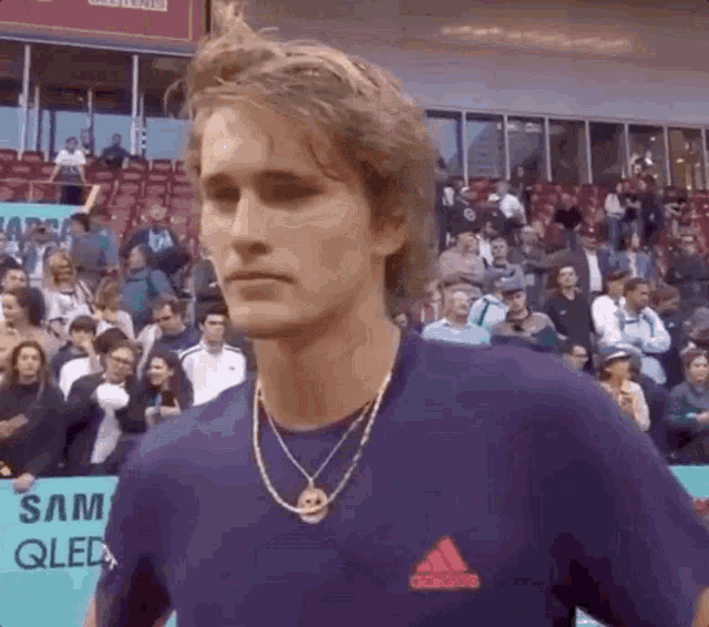 a man wearing a purple adidas shirt and a necklace is standing in front of a crowd .