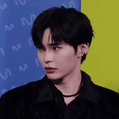 a young man wearing a black shirt and a necklace stands in front of a wall with the letter m on it