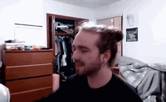 a man with long hair in a bun is sitting in a bedroom with a dresser and a bed .