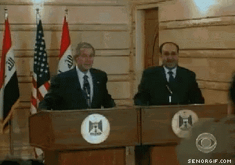a man in a suit and tie stands at a podium with flags behind him