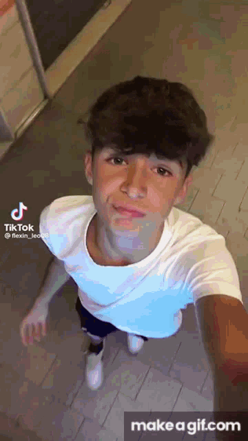 a young man in a white shirt is taking a selfie on a tile floor .