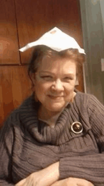 a woman wearing a sweater and a brooch is wearing a paper towel hat on her head .