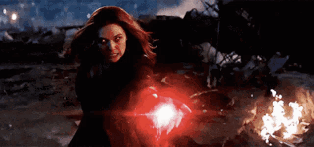 scarlet witch is holding a red light in her hand while standing next to a fire .
