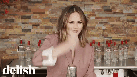 a woman in a pink suit is shaking a cocktail in front of a brick wall .