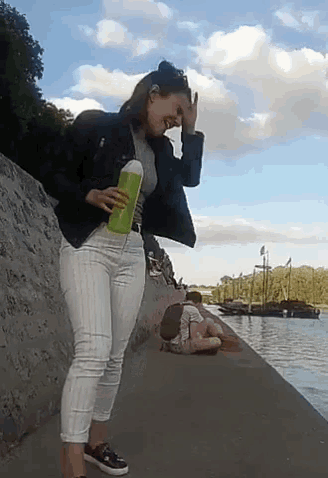 a woman in a black jacket and white pants is holding a green bottle while standing next to a body of water .