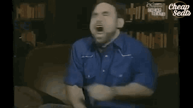a man in a blue shirt is sitting on a couch with his mouth open and screaming .