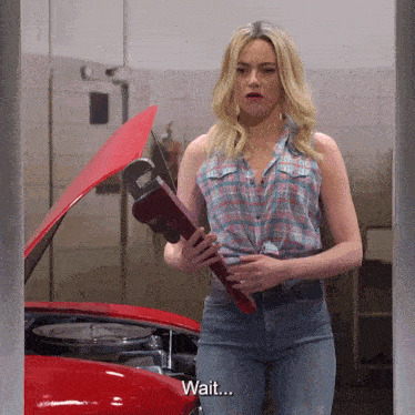 a woman in a plaid shirt holds a wrench in front of a red car and says wait