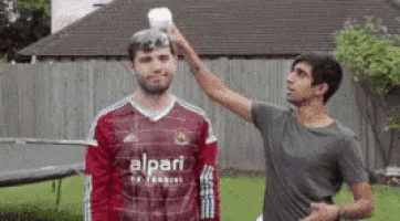 a man in a red alpari shirt is getting a bucket of water poured on his head .