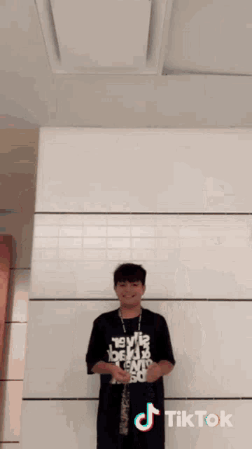 a boy is standing in front of a white wall with a tiktok logo on the bottom right
