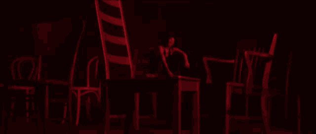 a woman sits at a table in a dark room surrounded by red chairs
