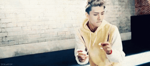 a young man in a yellow hoodie is eating something with chopsticks while sitting in front of a brick wall .