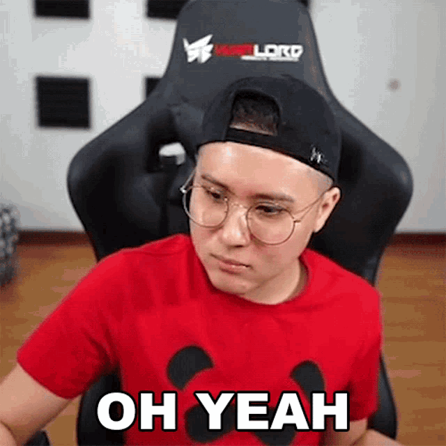 a man wearing glasses and a hat is sitting in a chair with the words oh yeah written on his shirt .