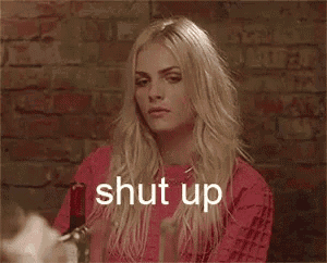a woman in a red shirt is sitting at a table with a bottle of wine and the word shut up .