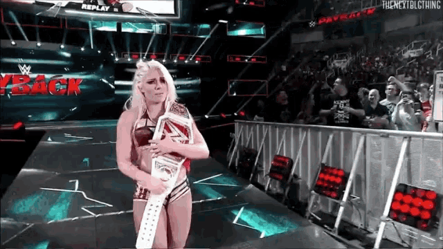 a female wrestler is holding a wrestling championship belt in front of a crowd and a sign that says replay