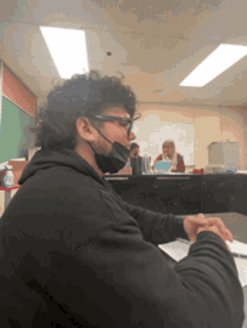 a man wearing a black mask sits at a desk in a classroom