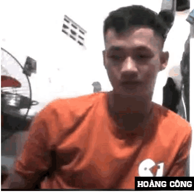 a man in an orange shirt is sitting in front of a white wall with the name hoàng công on the bottom