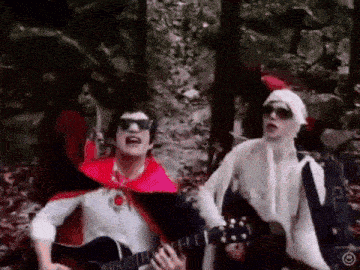a man in a red cape is playing a guitar next to another man