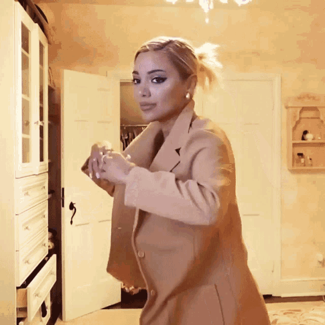 a woman wearing a tan coat is standing in a room