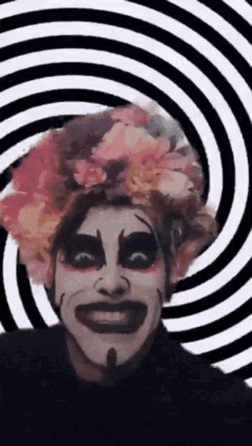 a clown with a flower crown on his head is smiling in front of a hypnotic spiral .