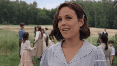 a woman in a blue shirt is smiling in front of a group of children in a field sponsored by hallmark