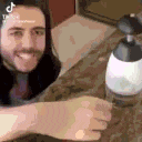 a man with long hair is sitting at a table with a soap dispenser in front of him .