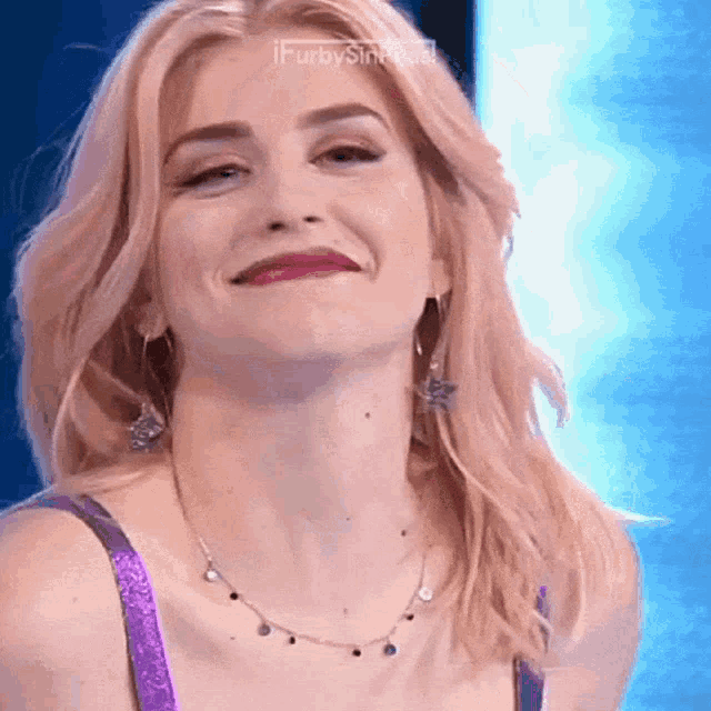 a woman wearing a purple bra and a necklace smiles for the camera with a blue background
