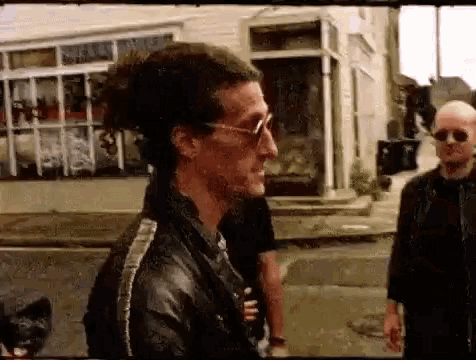 a man wearing sunglasses and a leather jacket is standing on a sidewalk next to a man wearing sunglasses .