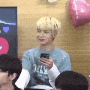 a man with blonde hair is holding a cell phone in his hand while sitting in a room with other people .