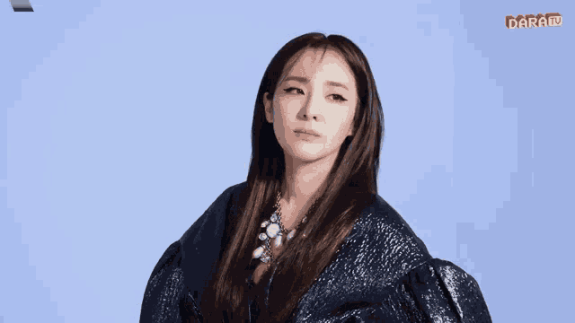 a woman in a sequined jacket stands in front of a blue background with the word darai on the bottom left